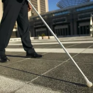 نمای نیمه پایینی فردی با شلوار و کفش مشکی که عصای سفیدی را به سمت بیرون در یک میدان سنگی در دست دارد