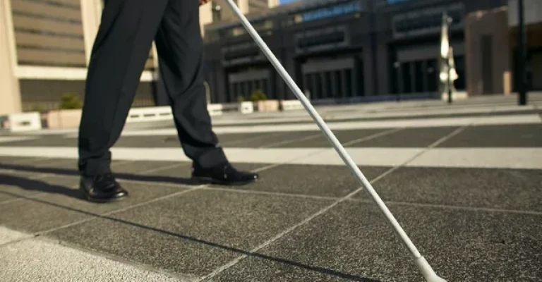 نمای نیمه پایینی فردی با شلوار و کفش مشکی که عصای سفیدی را به سمت بیرون در یک میدان سنگی در دست دارد